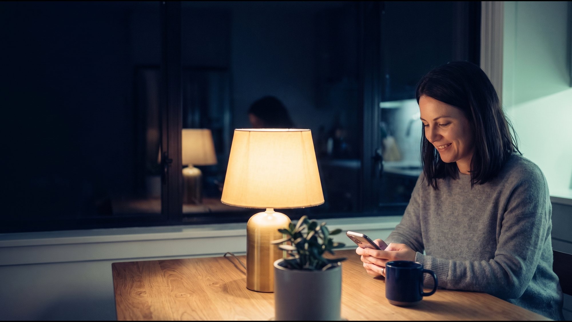 A woman at a kitchen table at night, typing a message on her phone.