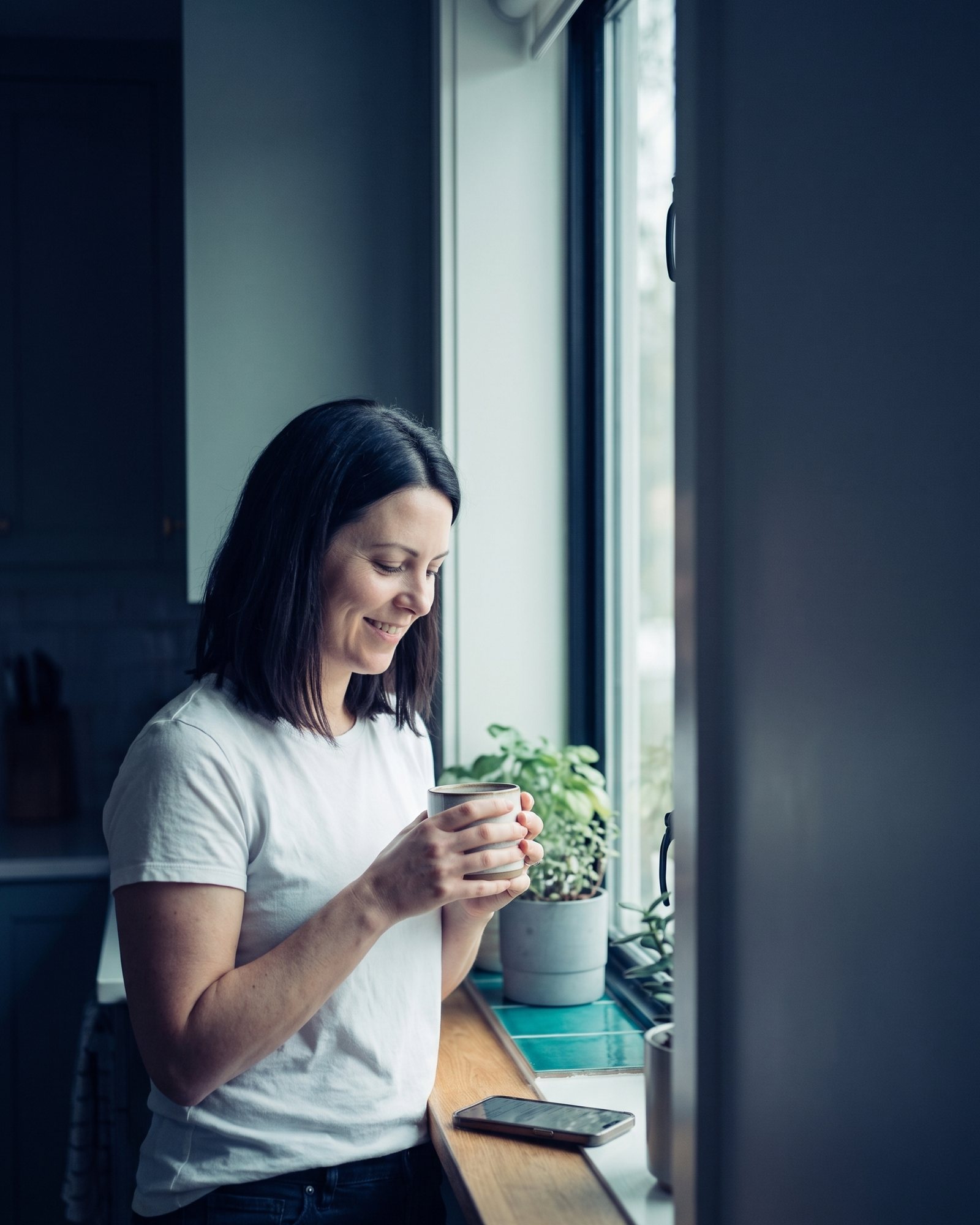 The same woman at a bright kitchen window in the morning, holding a mug.